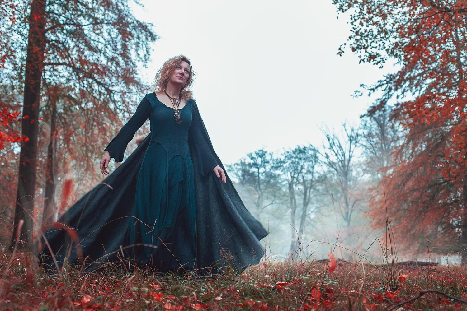 A woman in a flowing black dress with a long black coat walking through an autumn forest with red-orange leaves on the trees and ground.