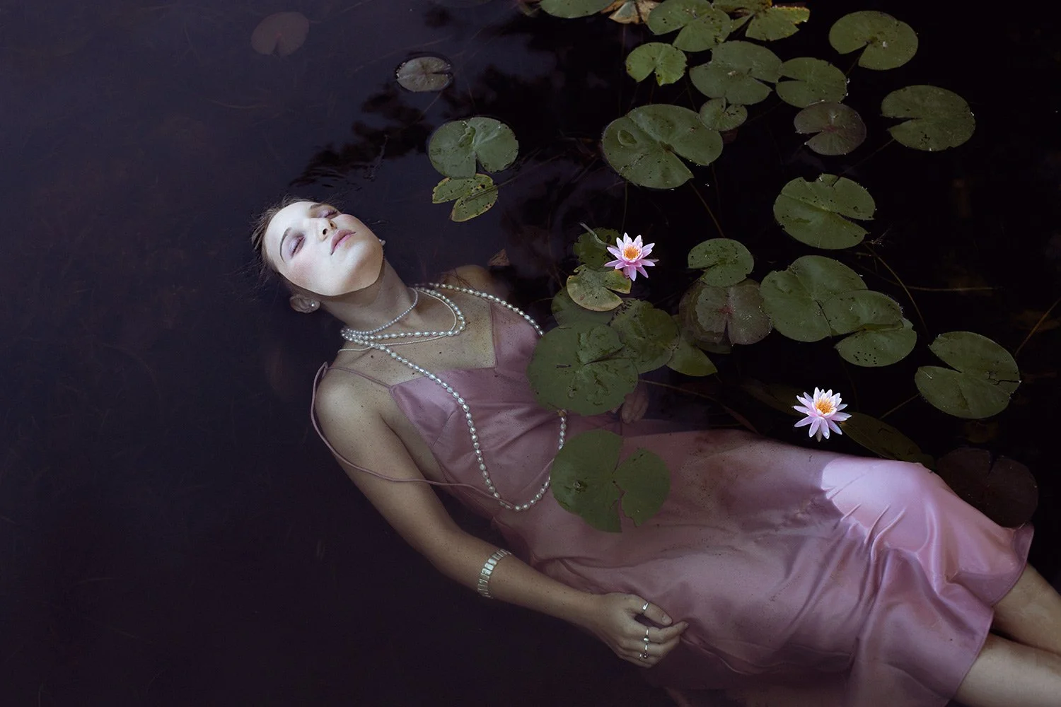 A woman wearing a pink satin dress and pearl jewelry lying in dark water among lily pads and pink water lilies, with her eyes closed.
