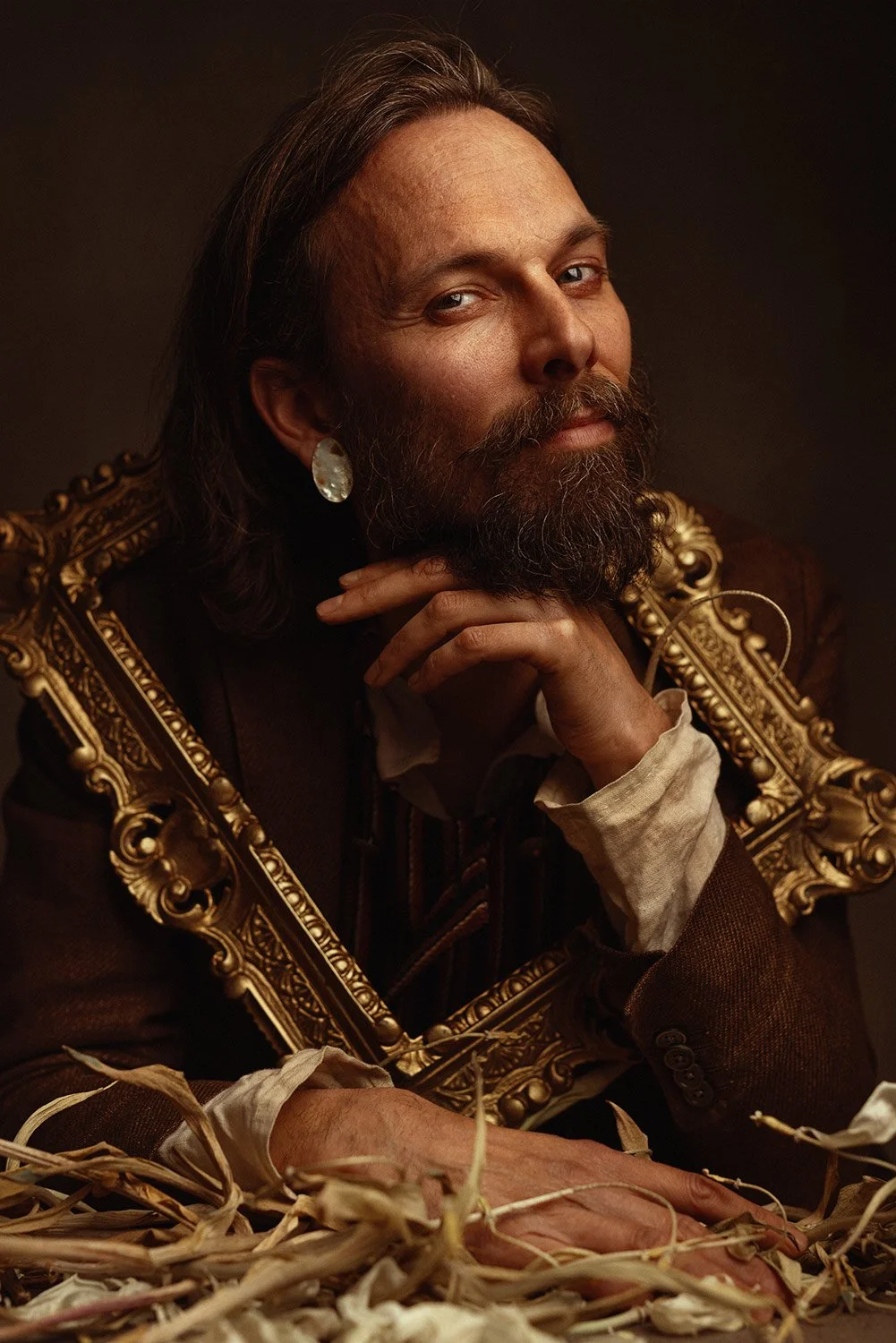 Portrait of a man with long hair and a beard, wearing a large ornate gold frame around his shoulders, a dark suit, white shirt, and an earring, sitting with his hand resting on his chin among dried plants.