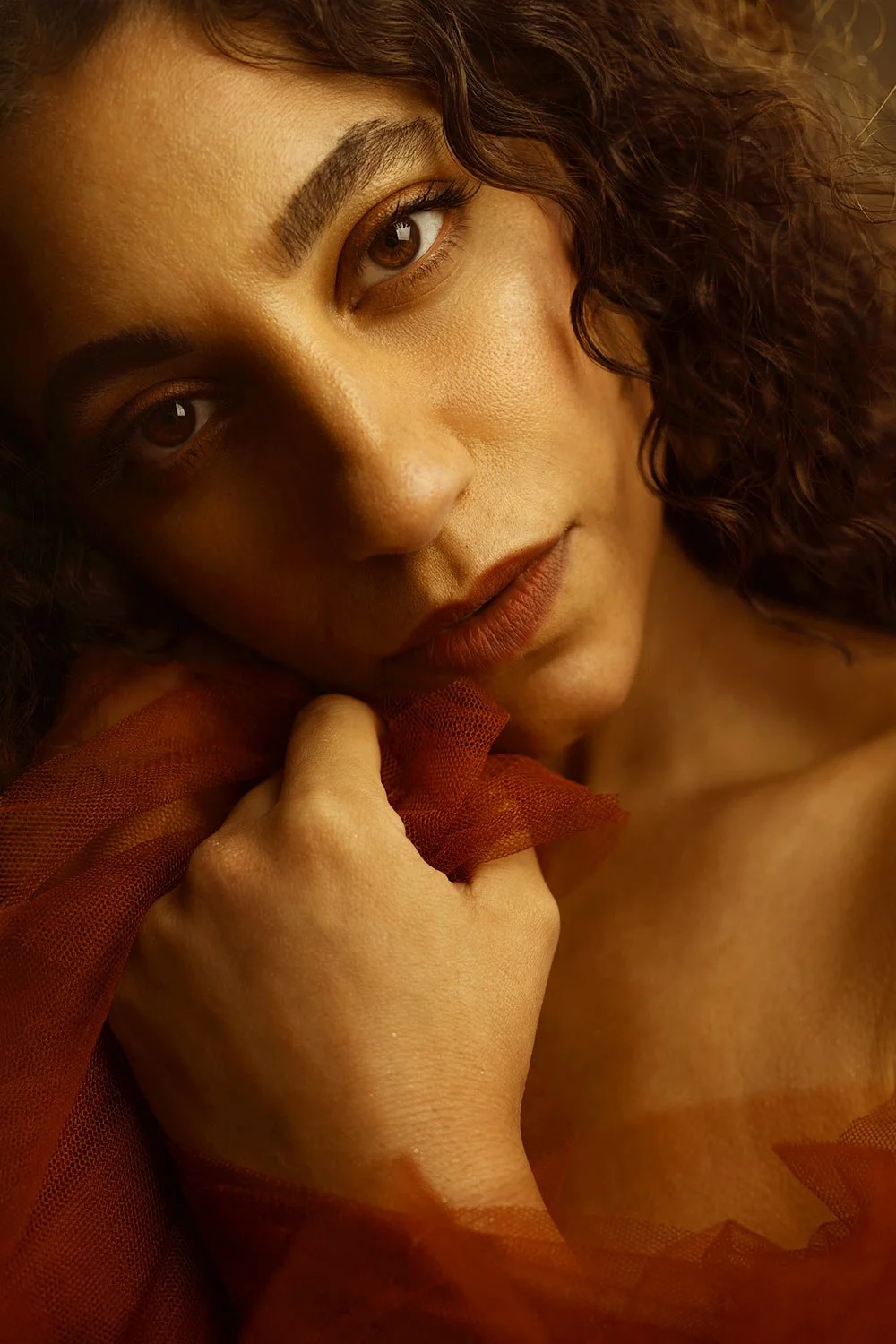 Close-up of a woman with curly hair holding a red sheer fabric near her face, looking directly at the camera with warm lighting.
