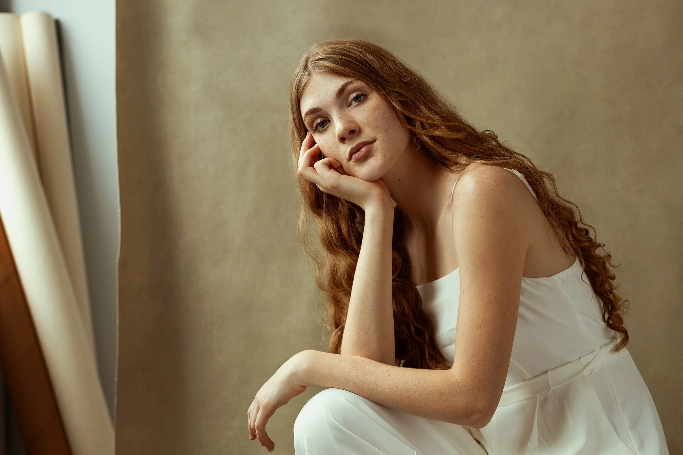 A young woman with long blond hair and fair skin wearing a white jumper, sitting with her elbow resting on her knee, resting her face on her hand, looking at the camera in natural light.