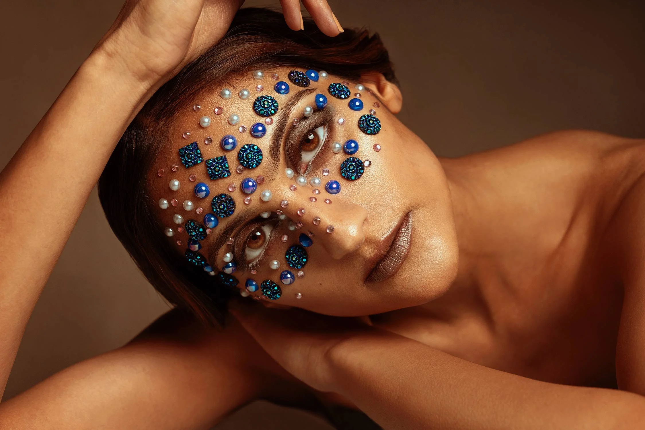 A woman with short brown hair and brown eyes adorned with blue and white decorative stones and pearls on her face, resting her head on her arm against a brown background.