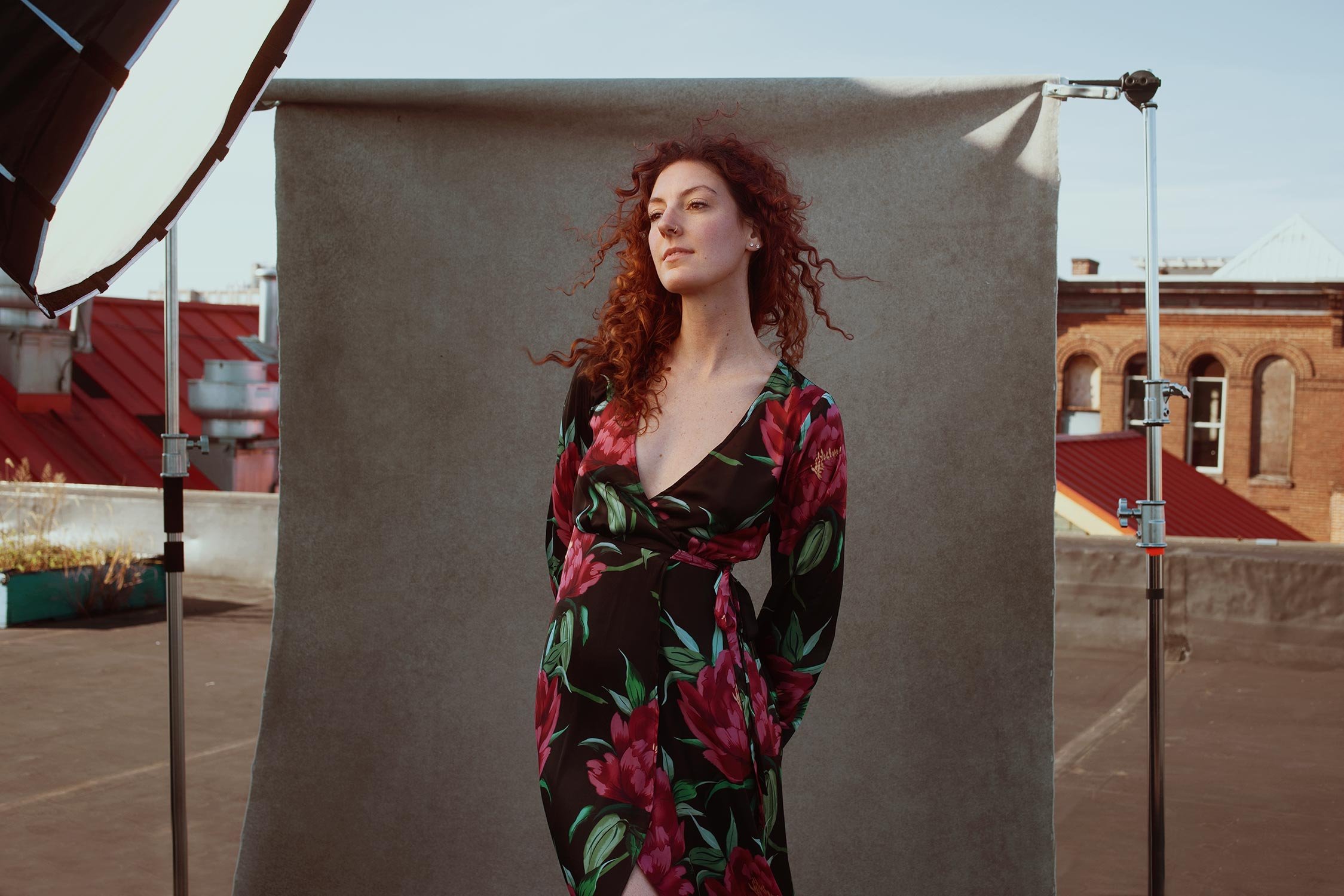A woman with red curly hair posing in front of a beige backdrop on a rooftop with red brick buildings in the background.