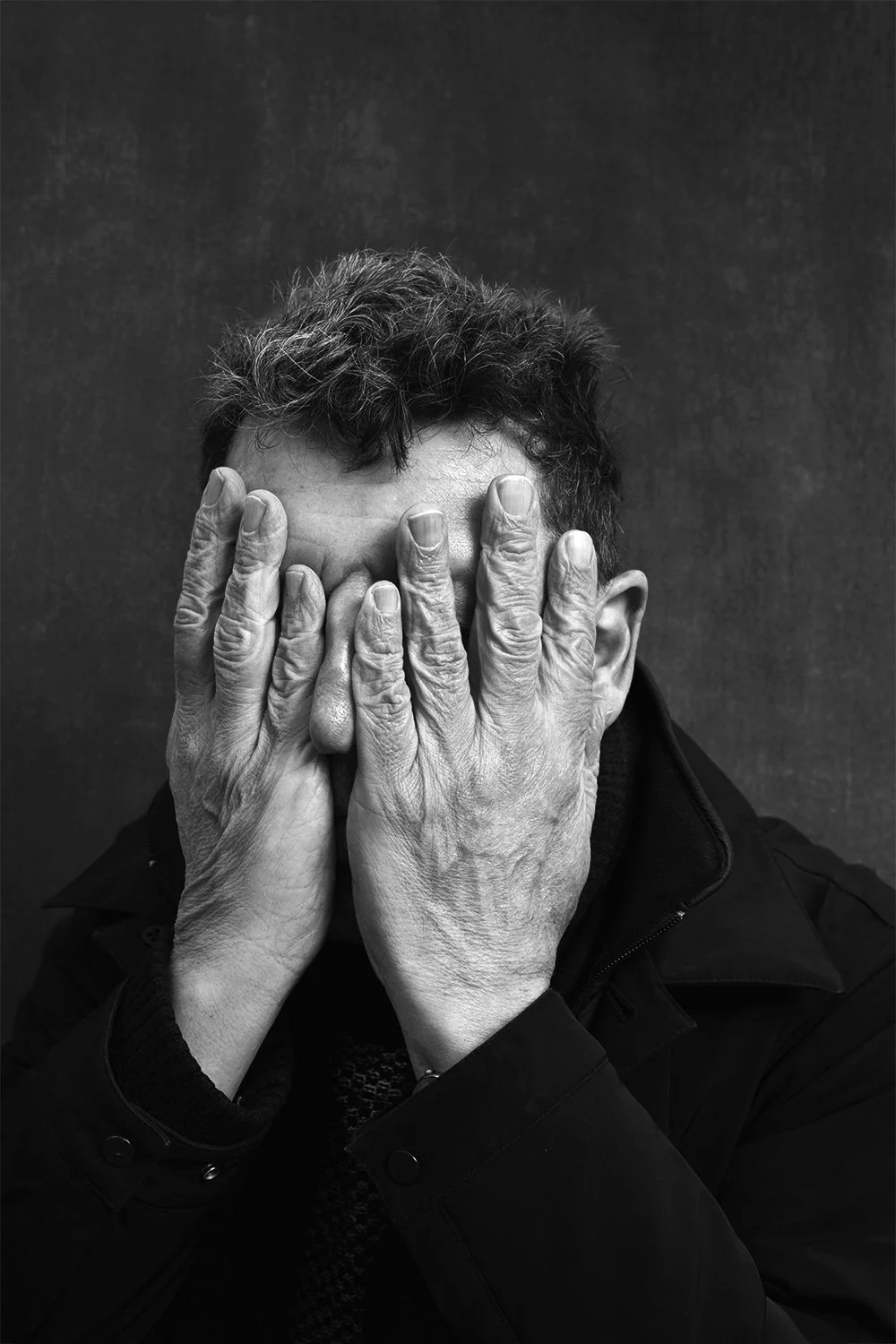 A black and white photograph of an elderly man covering his face with both hands, with his fingers spread apart, against a dark background.