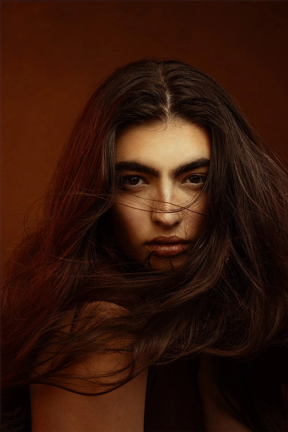 Close-up of a woman with flowing dark hair partially covering her face against a brown background.