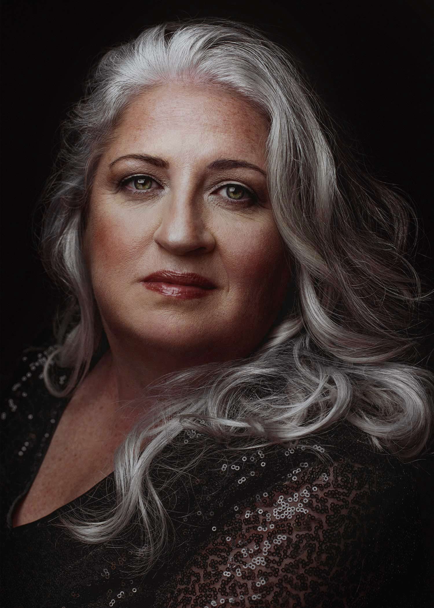 A portrait of a mature woman with long, wavy, gray hair, wearing a black sequin top, against a dark background.