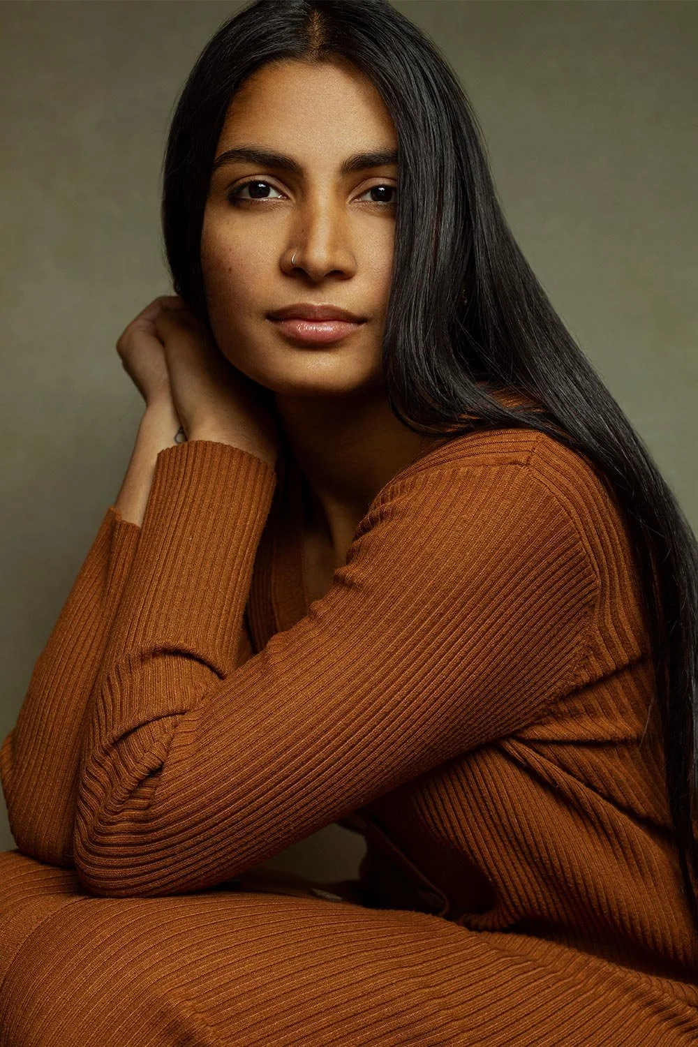 A young, Indian woman with long black hair, wearing a rust-colored ribbed long-sleeve top, looking at the camera with an intense expression.