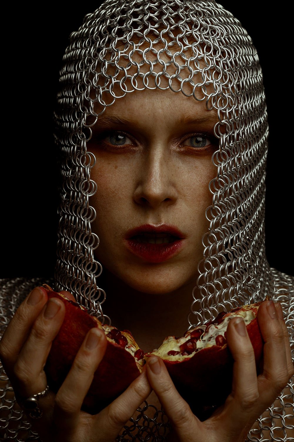 A woman wearing a chainmail hood holding a pomegranate half.