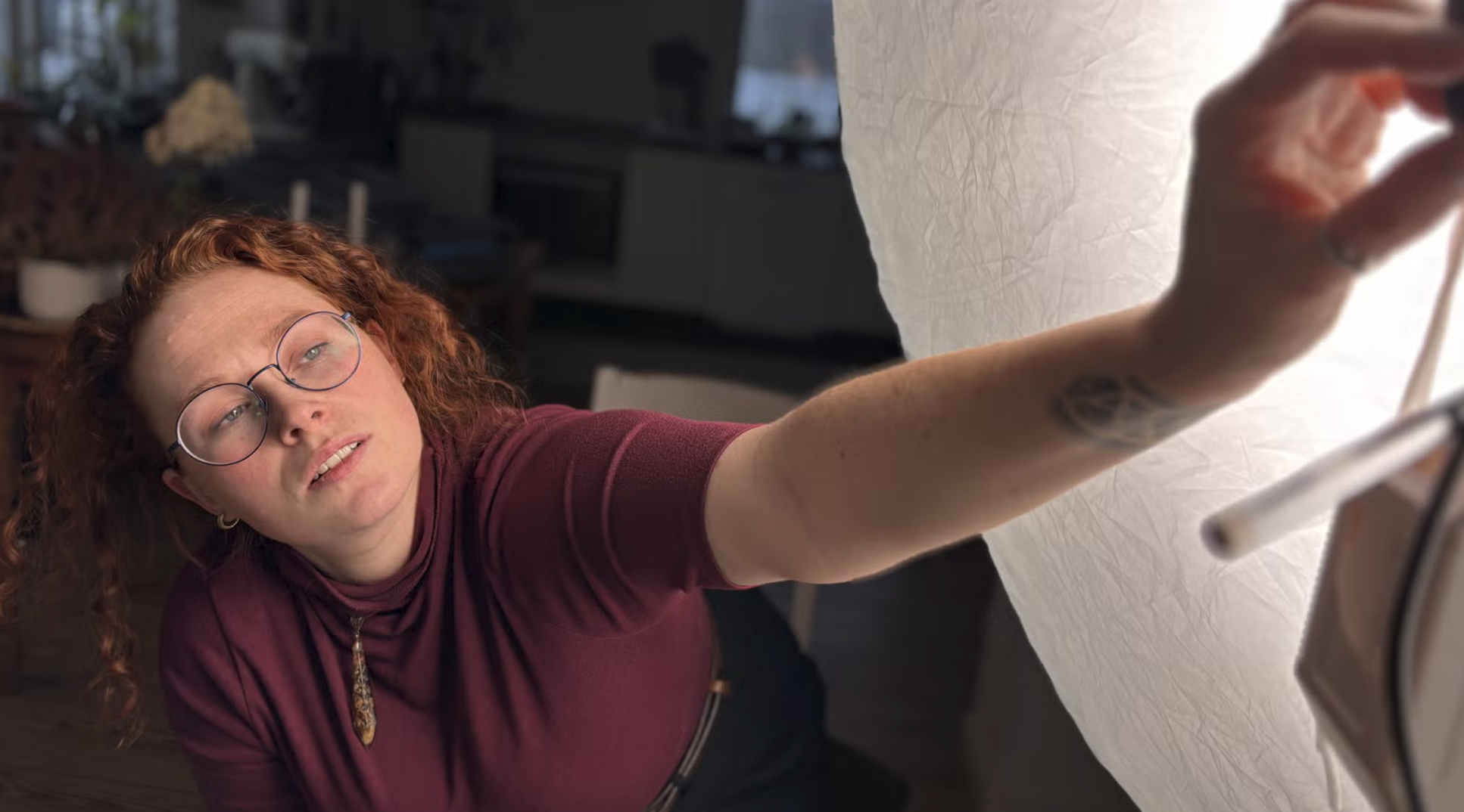 A woman with red hair, glasses, and a tattoo on her arm reaches out over a white surface, looking up at the camera.