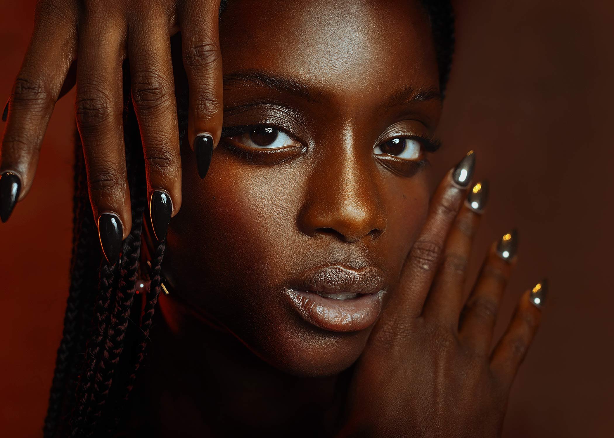 Close-up of a woman with dark skin, wearing metallic nail polish, touching her face with both hands, against a dark background.