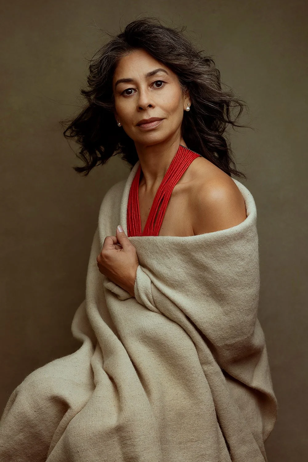 A woman with dark, wavy hair and pearl earrings, wearing a red top and beige shawl, looking at the camera against a plain background.