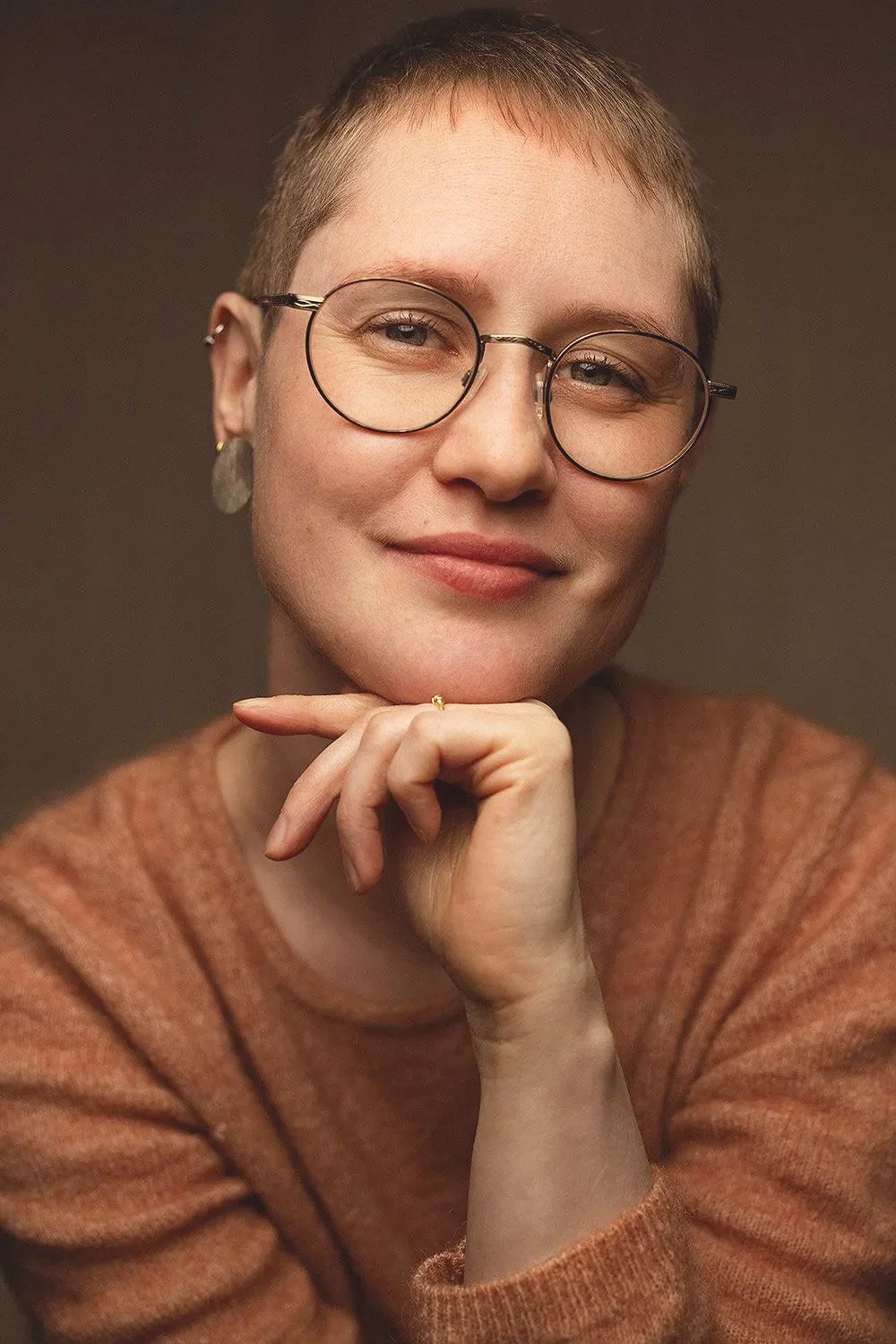 A woman with short light brown hair, glasses, and earrings, smiling gently and resting her chin on her hand, wearing a brown sweater.