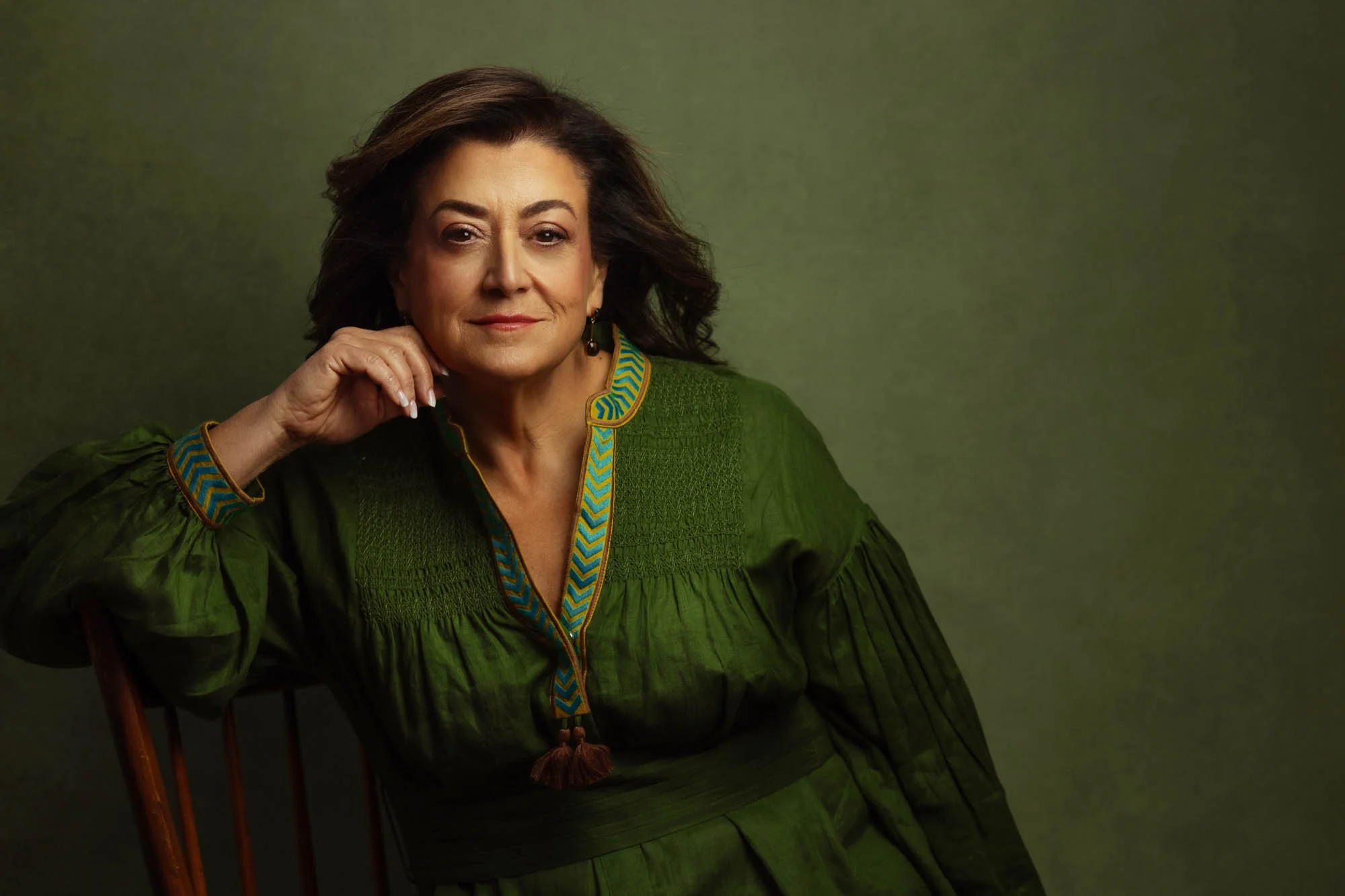 A portrait of an older woman with shoulder-length brown hair, wearing a green dress with a patterned trim and tassel, sitting on a wooden chair, looking at the camera with a confident expression, against a plain green background.