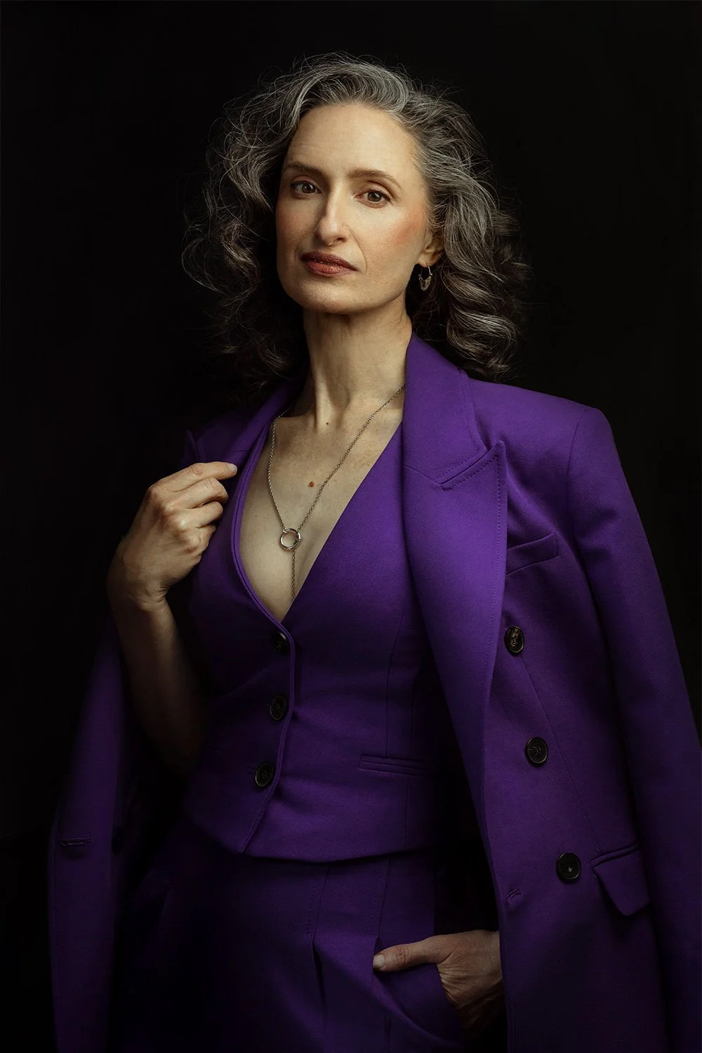 Portrait of a confident older woman with gray curly hair, wearing a purple suit and matching top, accessorized with a necklace and earrings, against a black background.