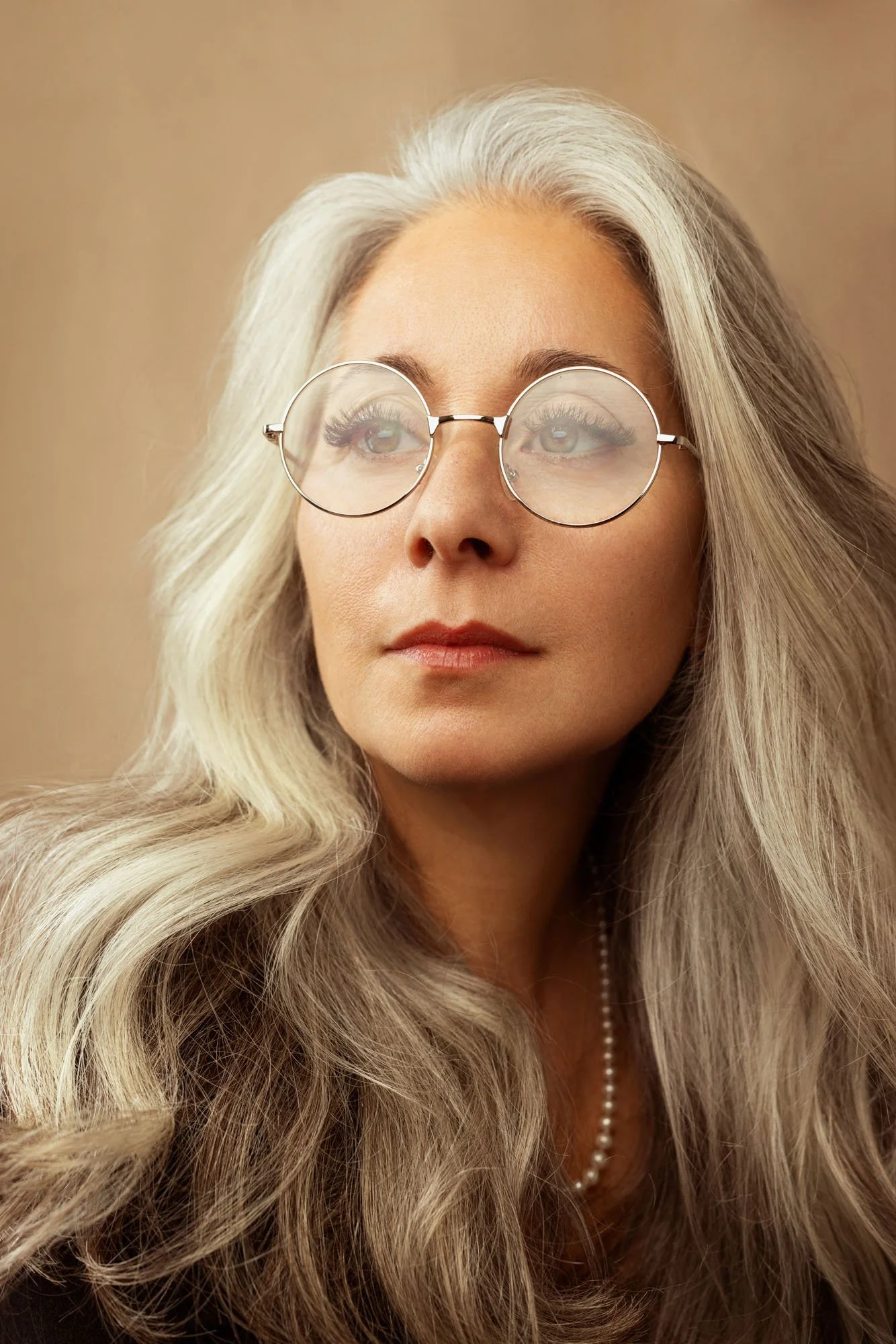 A woman with long, wavy, silver hair wearing round glasses with a gold frame, a small pearl necklace, and a black top, looking thoughtfully to the side with a beige background.