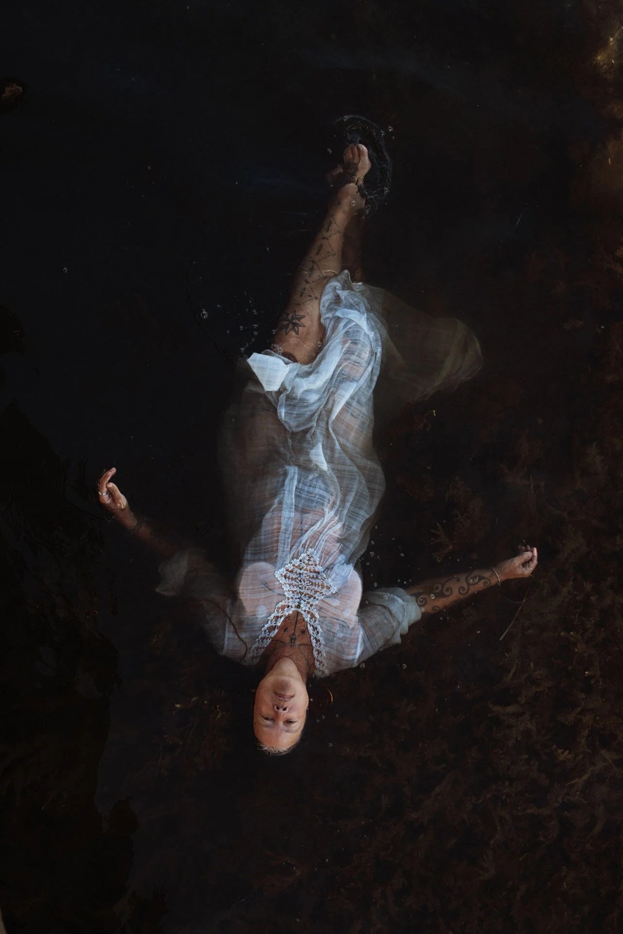 Person lying on a dark surface, partially submerged in water, wearing light-colored clothing with lace details, tattoos visible on arms and chest.