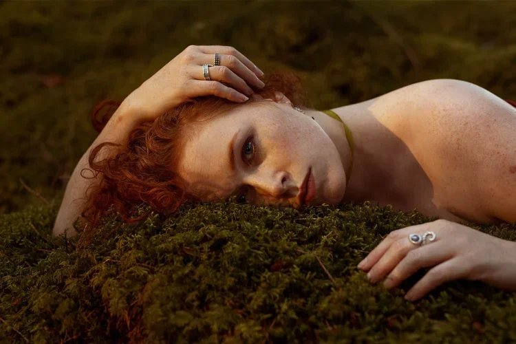 A woman with curly red hair lying on moss-covered ground, resting her head on her arm, looking at the camera.