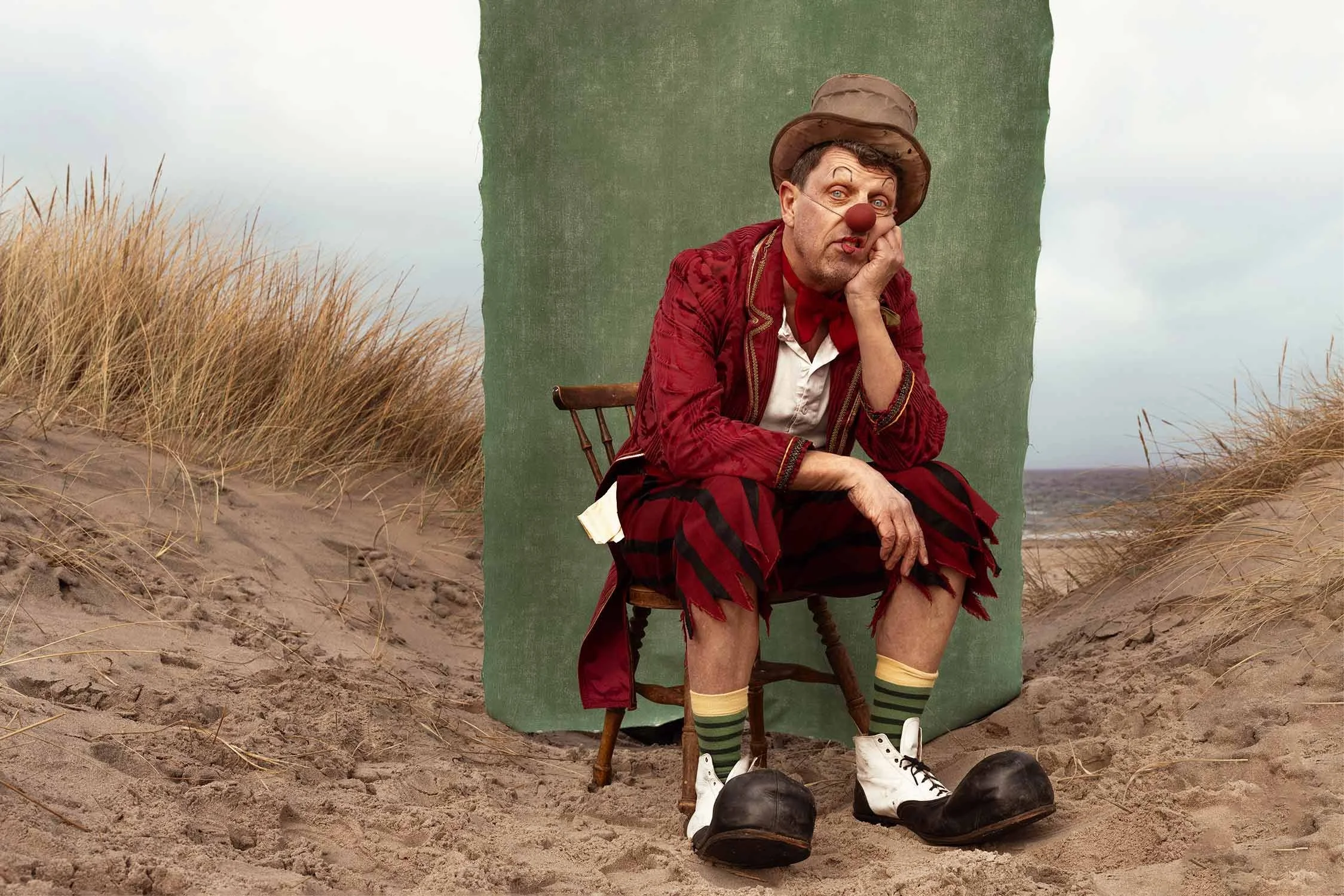A clown with a red nose, wearing a brown hat, red jacket, striped shorts, and oversized shoes, sitting on a wooden chair on a sandy beach with tall grass and a cloudy sky in the background.