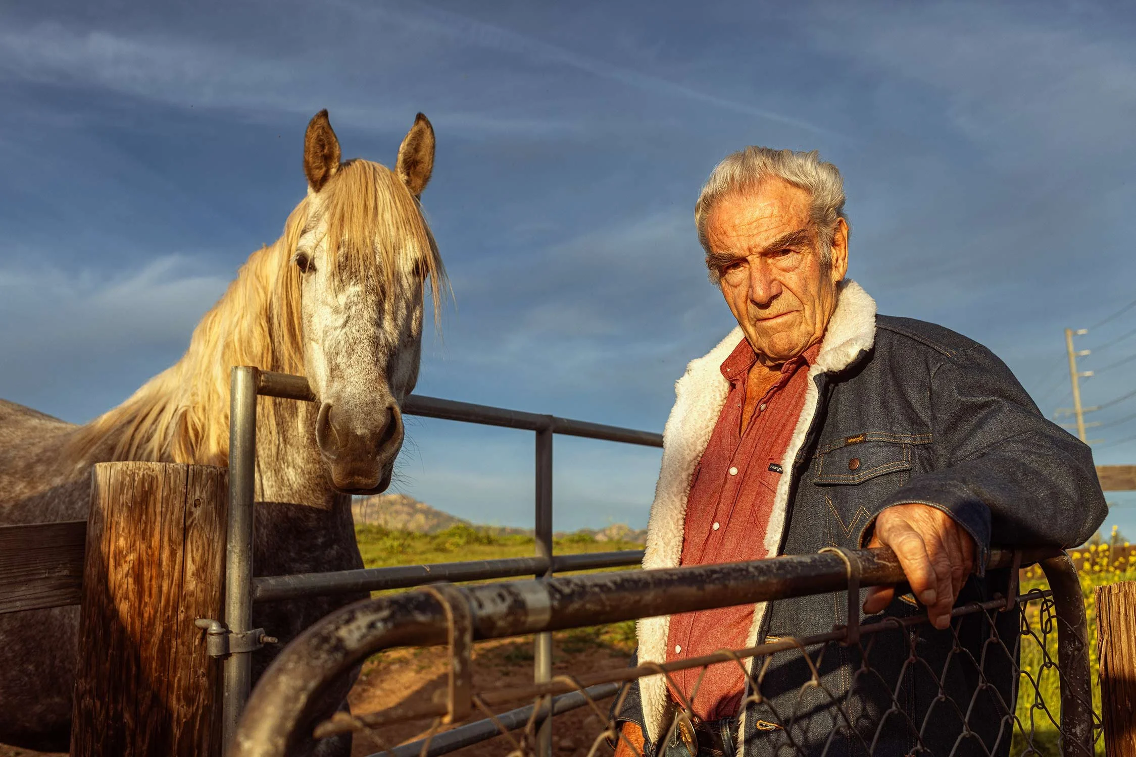 An older man leaning on a farm gate, standing next to a white horse with a long blonde mane, outdoors on a sunny day with a blue sky and distant hills in the background.