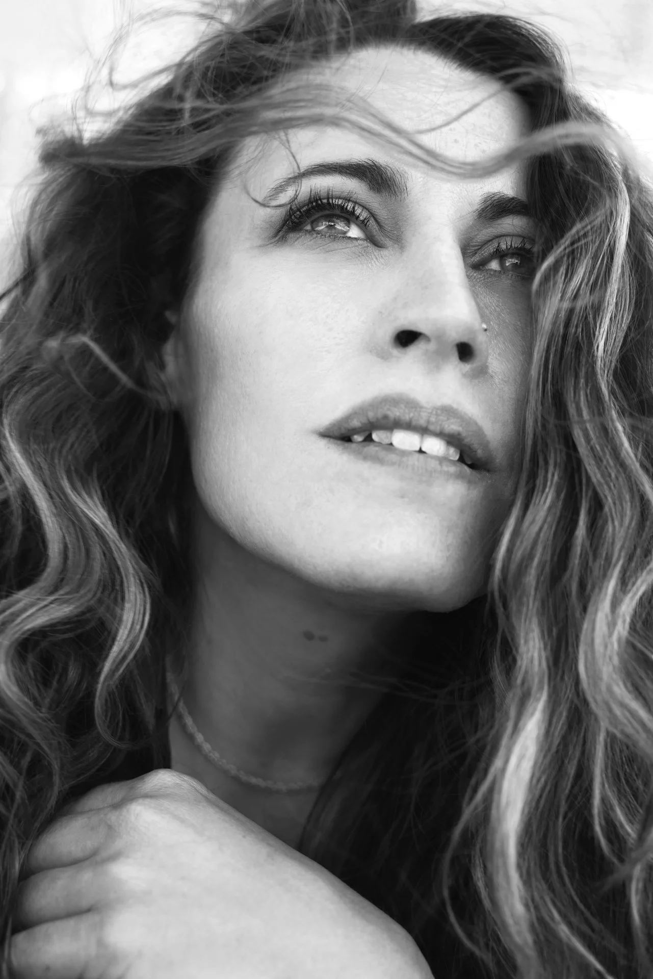 A close-up black and white portrait of a woman with long, wavy hair, looking upwards with a dreamy expression, her hand resting near her neck.