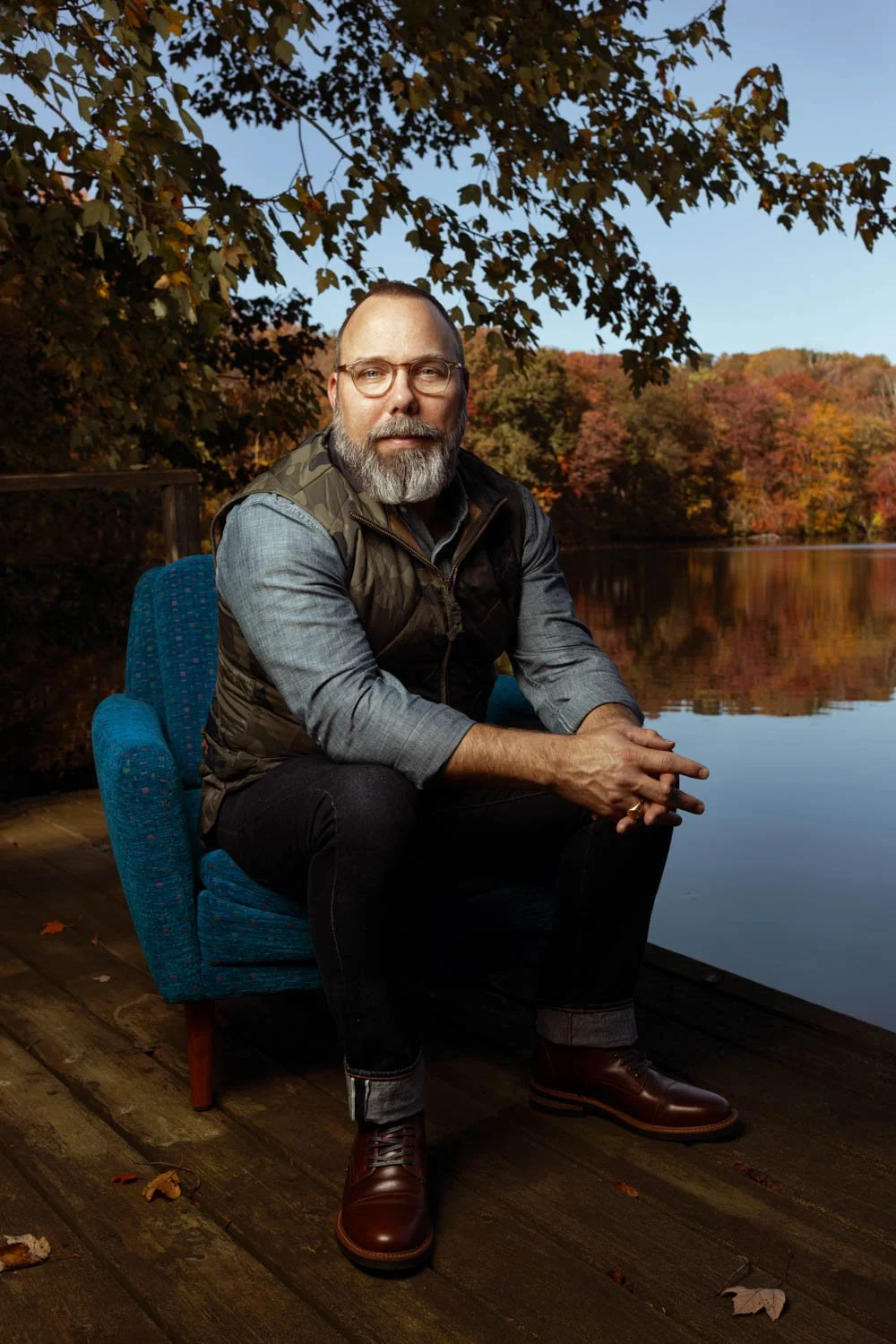 A man with glasses, beard, and gray hair sitting on a blue upholstered chair on a wooden dock by a lake, with fall foliage in the background.