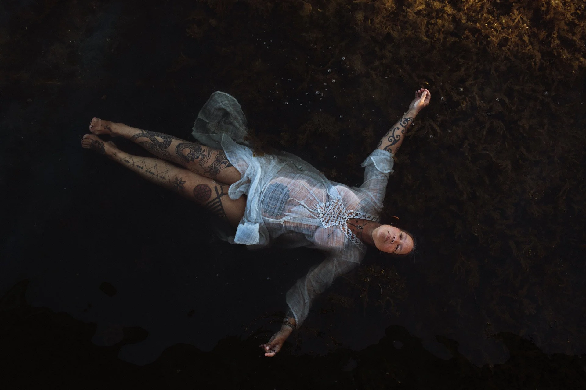 An older woman with full-body tattoos lies floating on water with dark aquatic plants beneath her, wearing a sheer, long-sleeved dress.