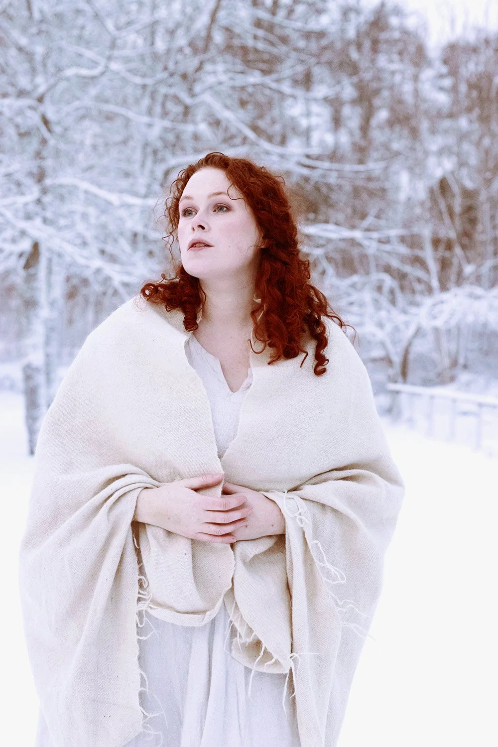 Woman with curly red hair standing outdoors in a snowy landscape, wearing a white cloak and looking to the side.