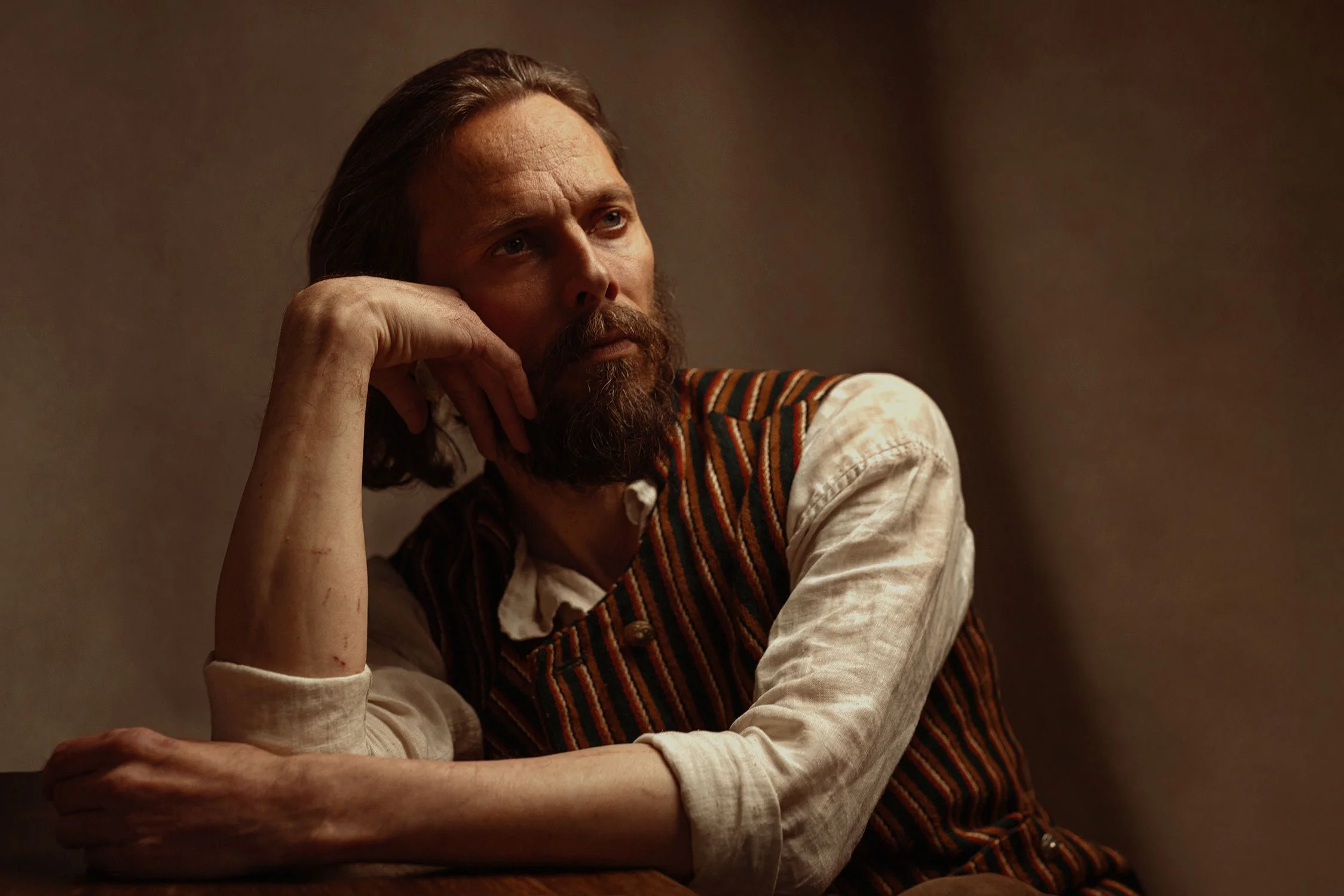 A man with long hair and a beard, wearing a vintage-style vest over a beige shirt, sits with his head resting on his hand, looking thoughtfully into the distance in a dimly lit room.