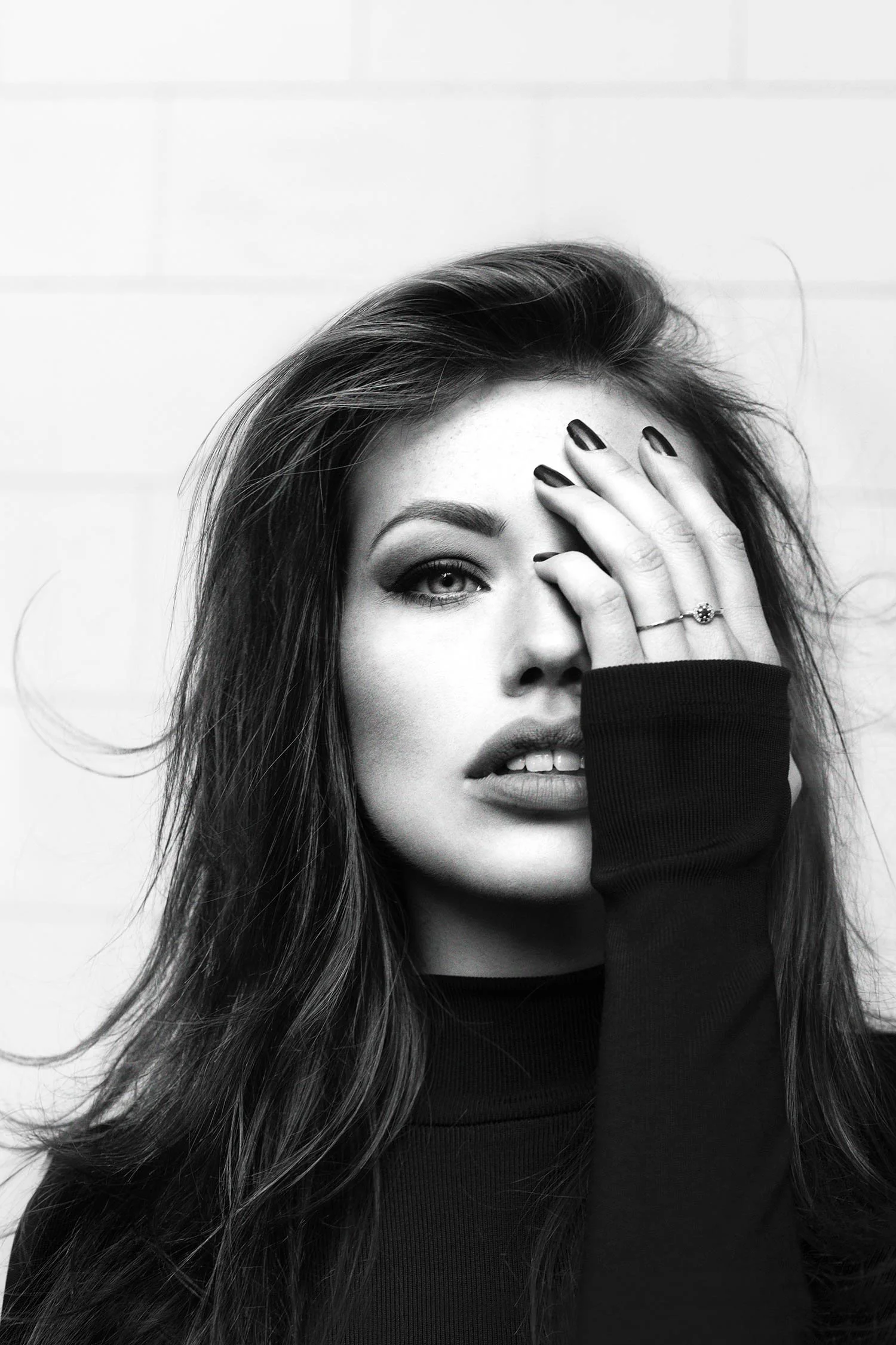 Black and white portrait of a woman with long hair, one eye visible, hand covering part of her face, wearing a ring and a long sleeve top.