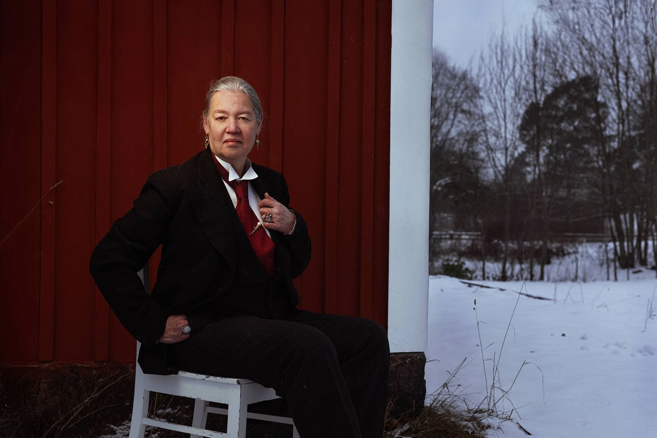 A woman with gray hair sitting on a white chair outside near a red wooden wall, on a winter day with snow-covered ground and leafless trees in the background.