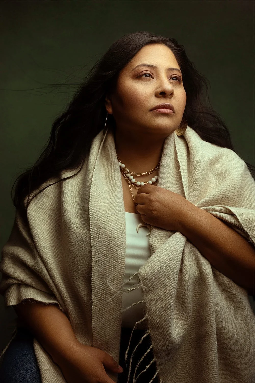 A woman with dark hair wearing a woven beige shawl, white top, pearl and gold jewelry, and gold earrings, looking contemplative against a dark green background.