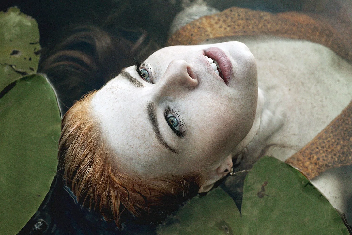 A woman with red hair and blue eyes lying on water among lily pads, looking up with a gentle expression.