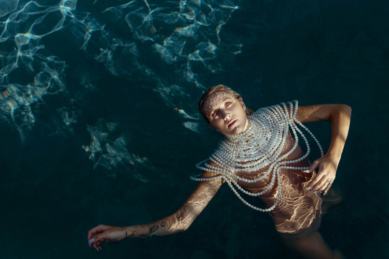 A woman floating in a dark pool, decorated with a pearl necklace and face jewelry, with sunlight creating reflections on her skin and water surface.