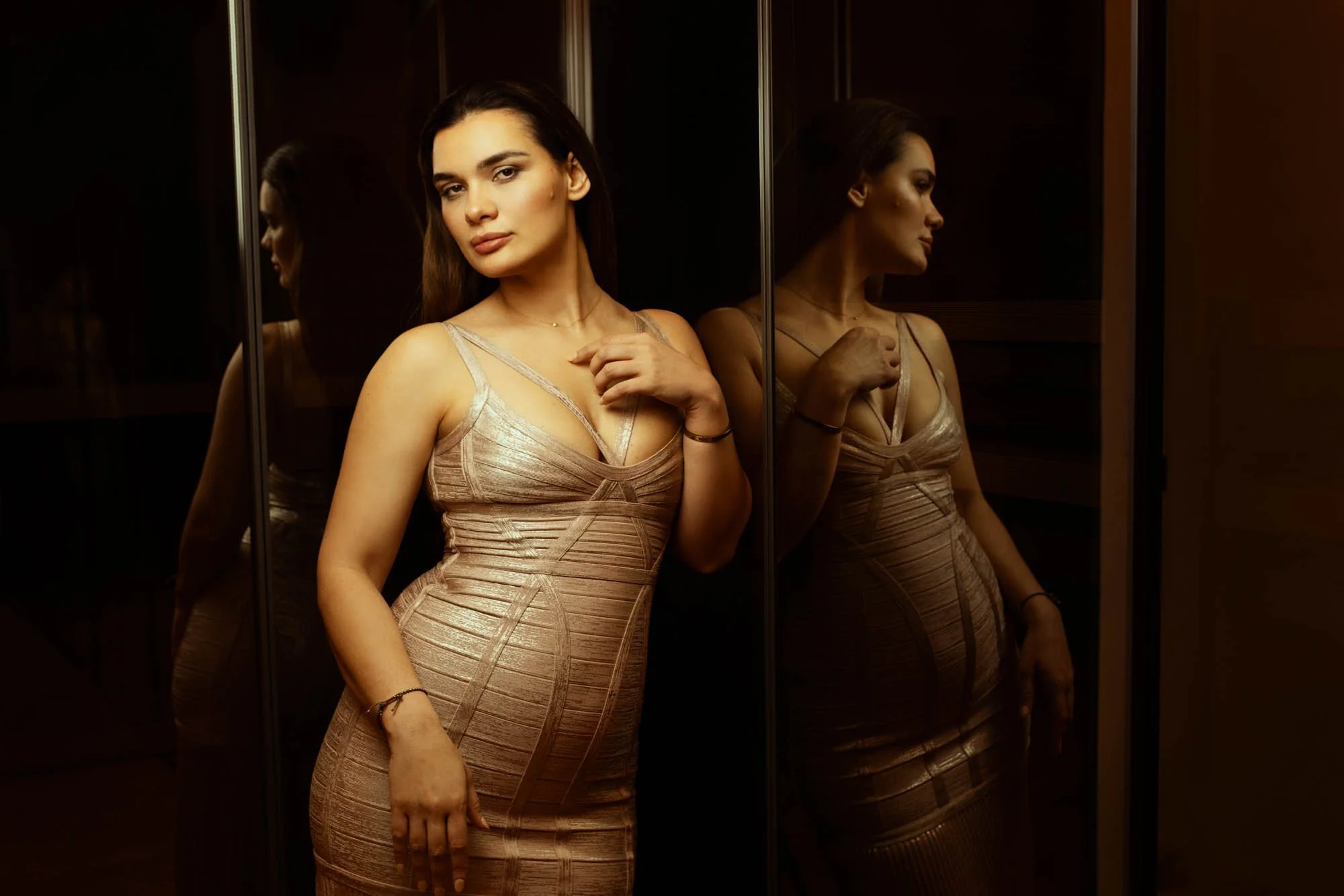 A young woman with brown hair and light skin standing in front of a mirror, wearing a form-fitting beige dress with thin straps, posing with one hand near her shoulder and the other on her thigh. Her reflection is visible in the mirror.