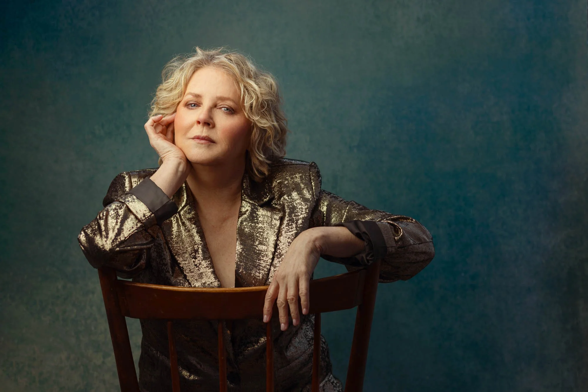 A middle-aged woman with blonde curly hair, wearing a gold and black patterned blazer, sitting on a wooden chair against a blue-green textured background, resting her head on her left hand, looking directly at the camera.
