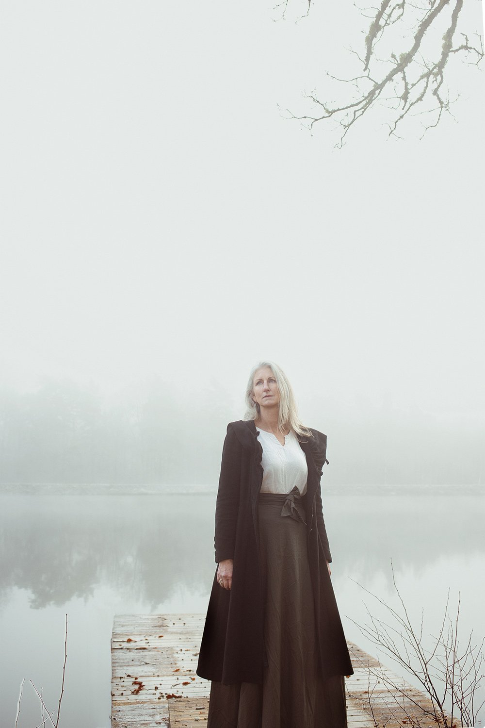 A woman with long gray hair standing on a wooden dock by a foggy lake, wearing a white blouse and a long dark skirt and coat.