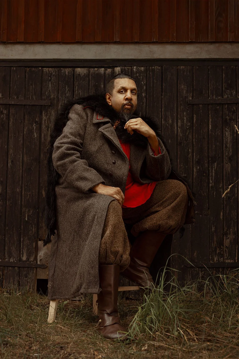 Man sitting on a wooden chair outdoors, wearing a brown coat, brown pants, and tall boots, with a dark beard and long hair, posing against a dark wooden background.