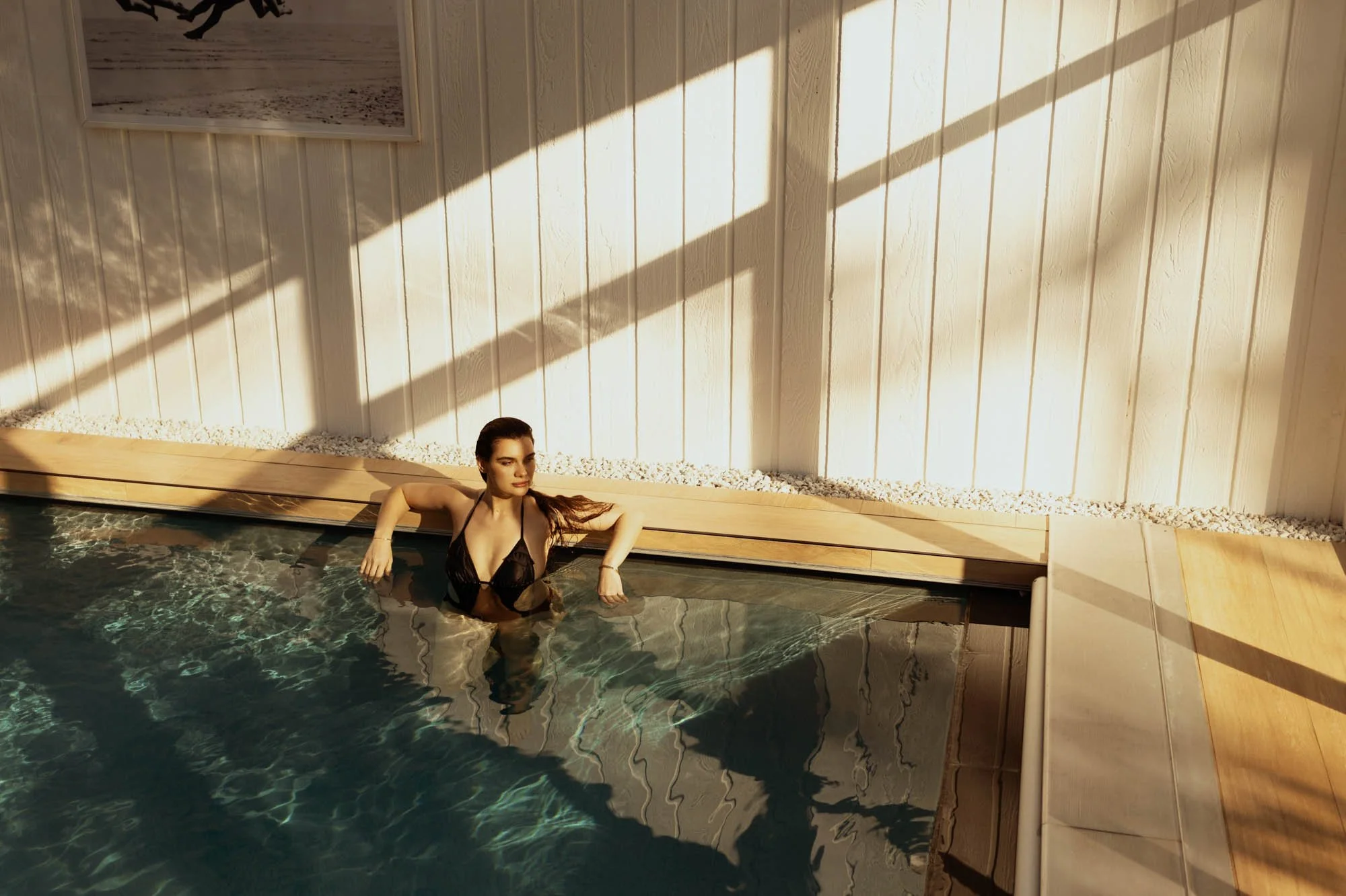 A woman in a black bikini relaxing in a pool with sunlight casting shadows on a wooden-paneled wall behind her.