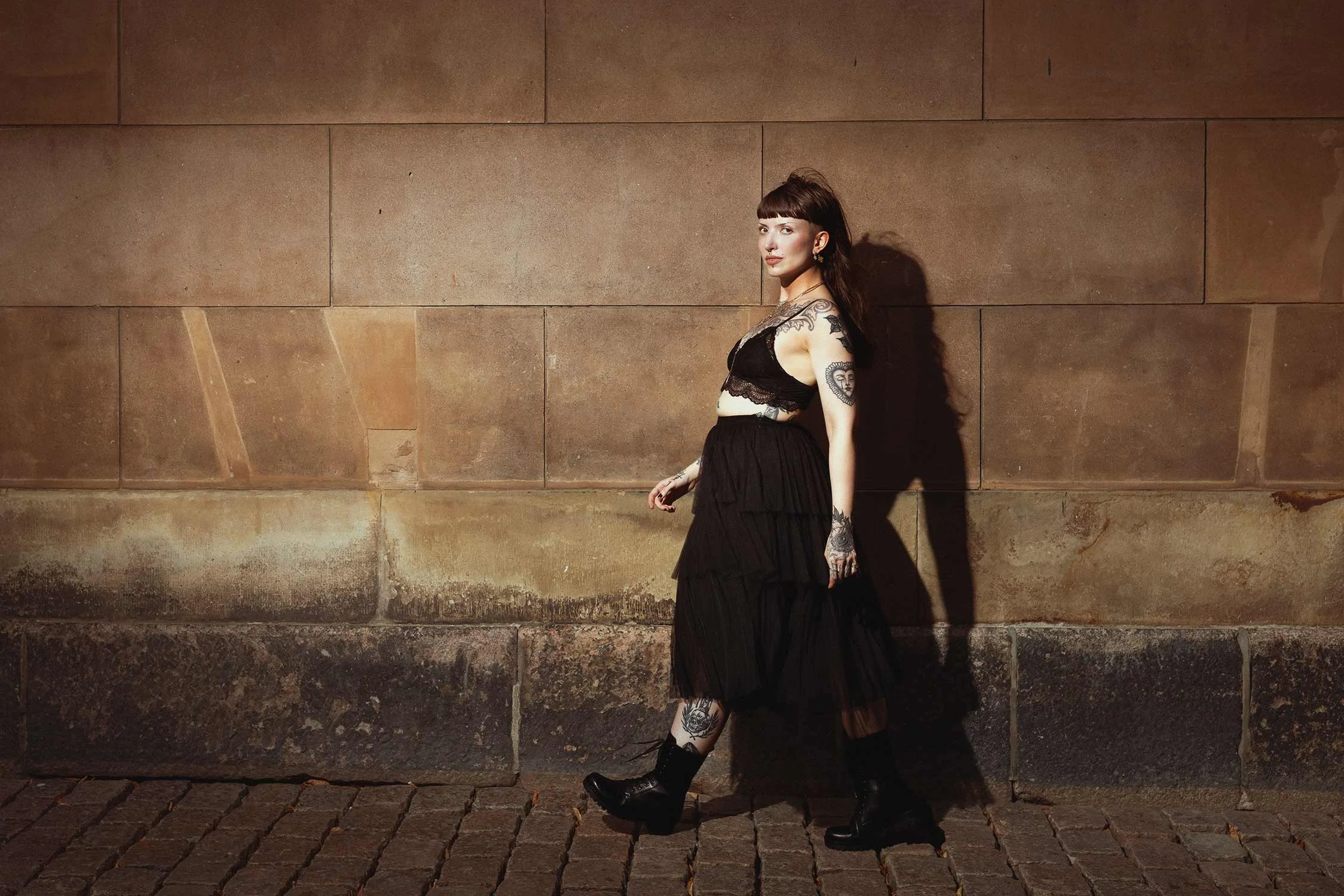 A woman with tattoos, dark brown hair, and a fringe hairstyle standing against a stone wall. She is wearing a black lace crop top, a layered black skirt, and black combat boots.