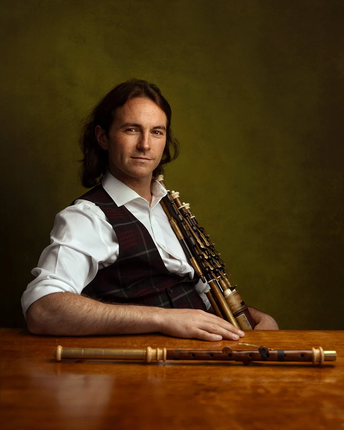 A man with long brown hair wearing a white dress shirt and a tartan vest, sitting at a wooden table with two musical instruments, a set of bagpipes draped over his shoulder and a flute lying on the table.