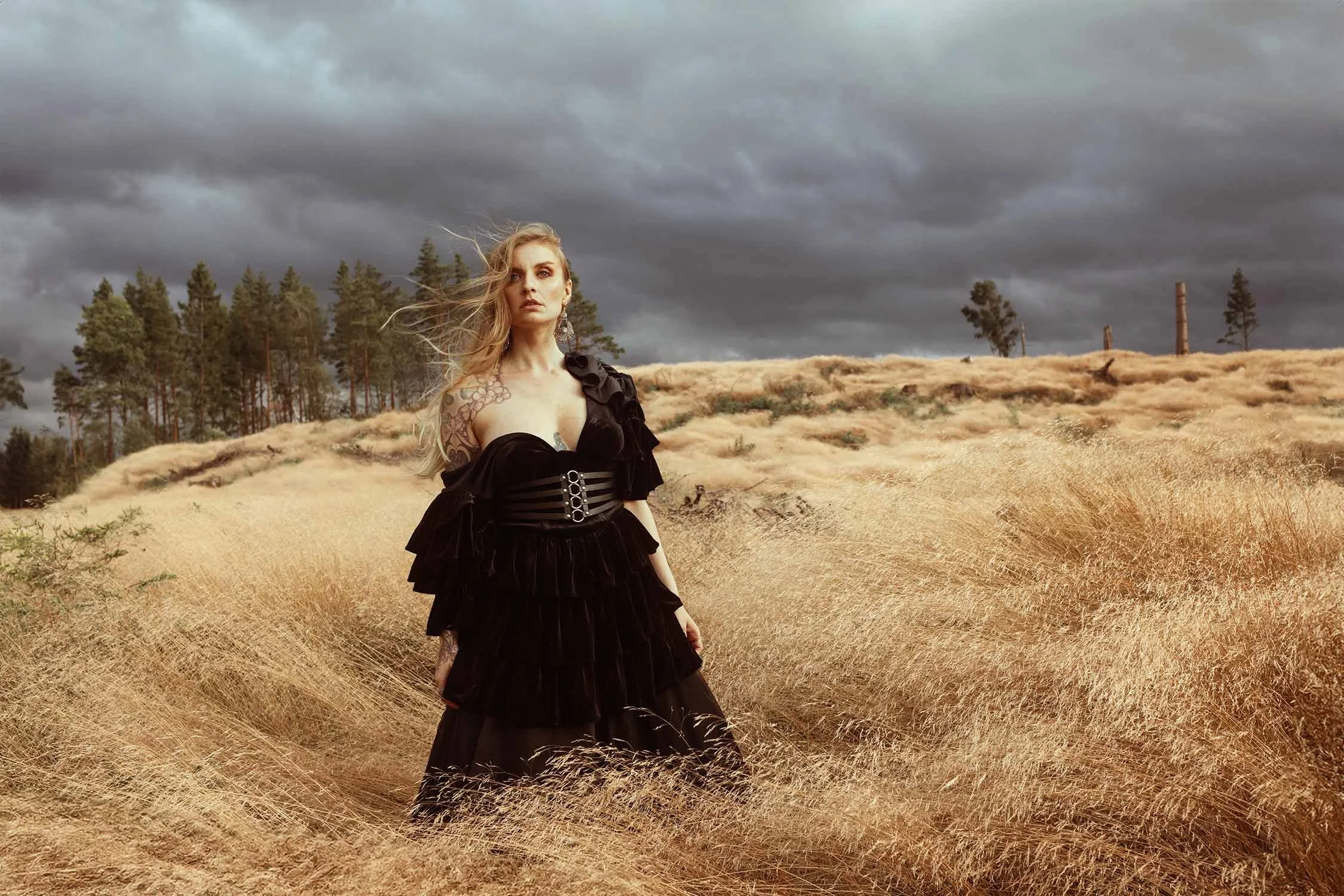 A woman wearing a black dress with ruffles and a corset style belt standing in a field of tall, dry grass under a cloudy sky with a forest and distant trees in the background.