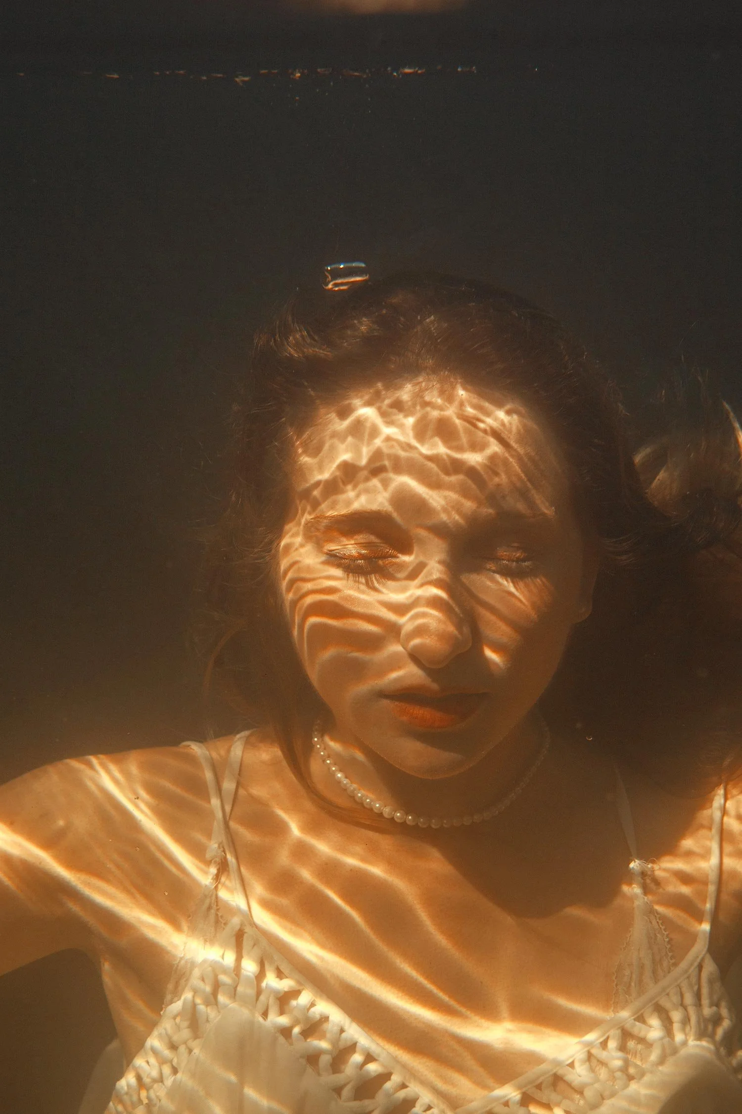 A woman with long dark hair and makeup floating in water, with sunlight creating rainbow-like patterns on her face and body.