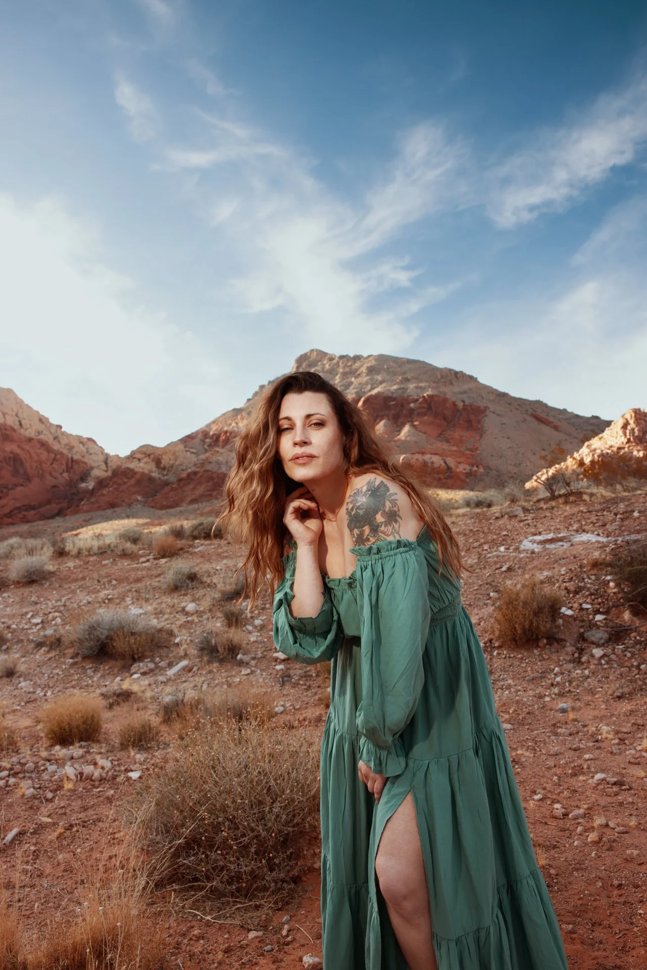A woman with long wavy hair and a tattoo on her left shoulder stands in a desert landscape with mountains in the background, wearing a green off-shoulder dress.