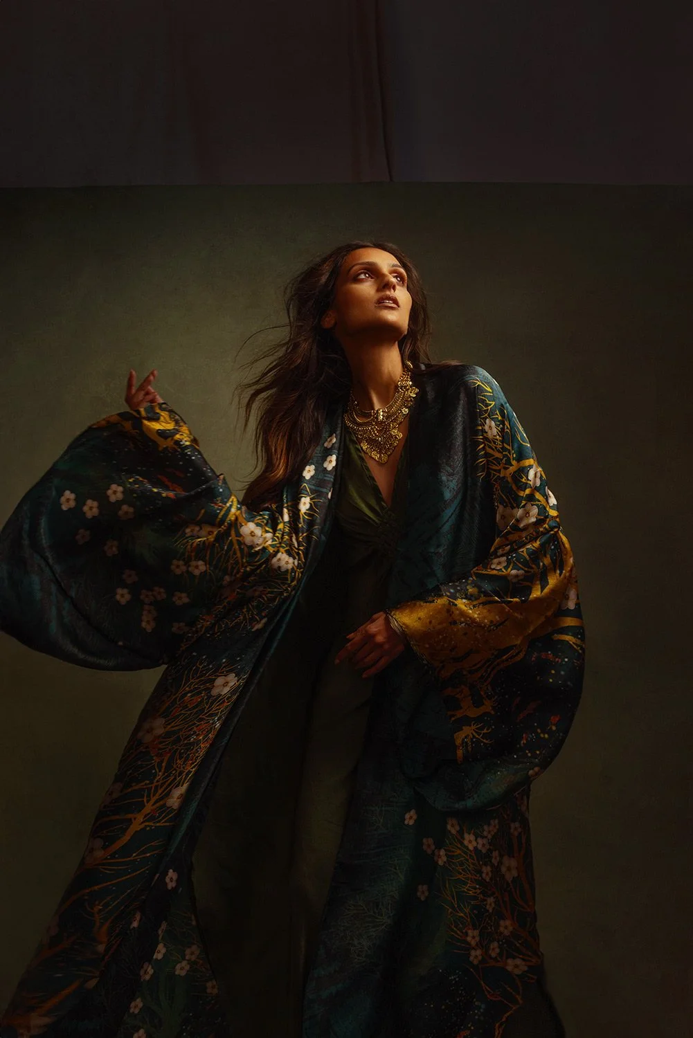 A woman with long dark hair wearing a dark green dress, insignia jewelry, and a colorful, patterned kimono with floral and abstract designs, standing against a plain background, looking upward with a confident expression.