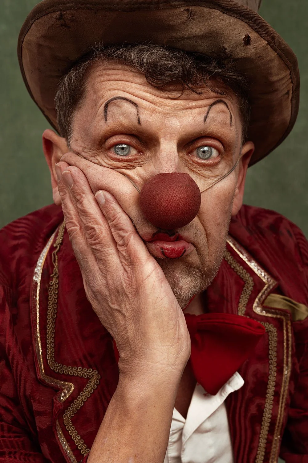 A person dressed as a circus clown with a red nose, face paint, and clown makeup, wearing a red velvet costume and a brown hat, with a green background.