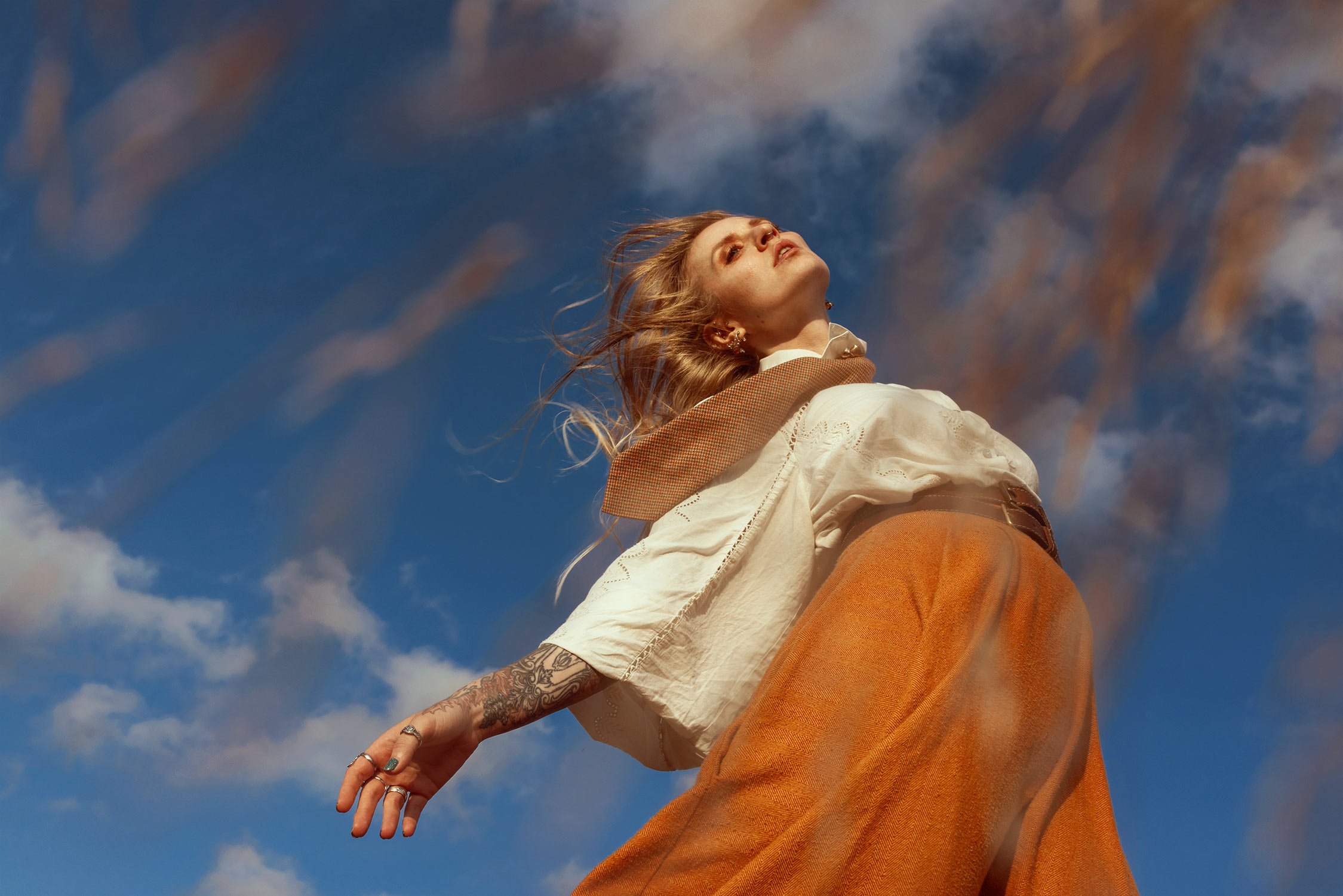 A woman with blonde hair and tattoos on her arm stands outdoors against a blue sky with scattered clouds, looking up with her hair blowing in the wind.