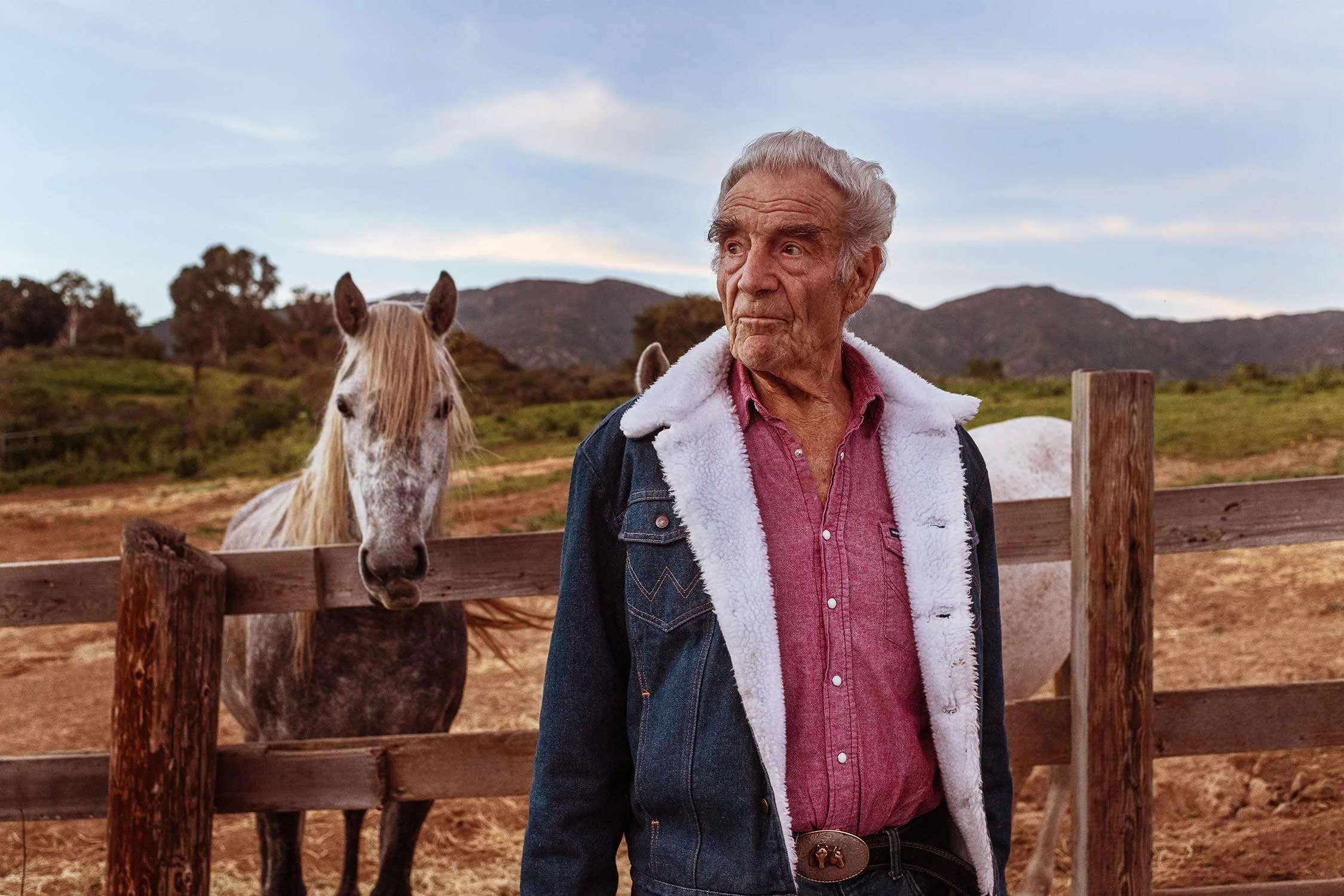 An elderly man standing outdoors near a wooden fence with two horses in the background, with hills and a partly cloudy sky.