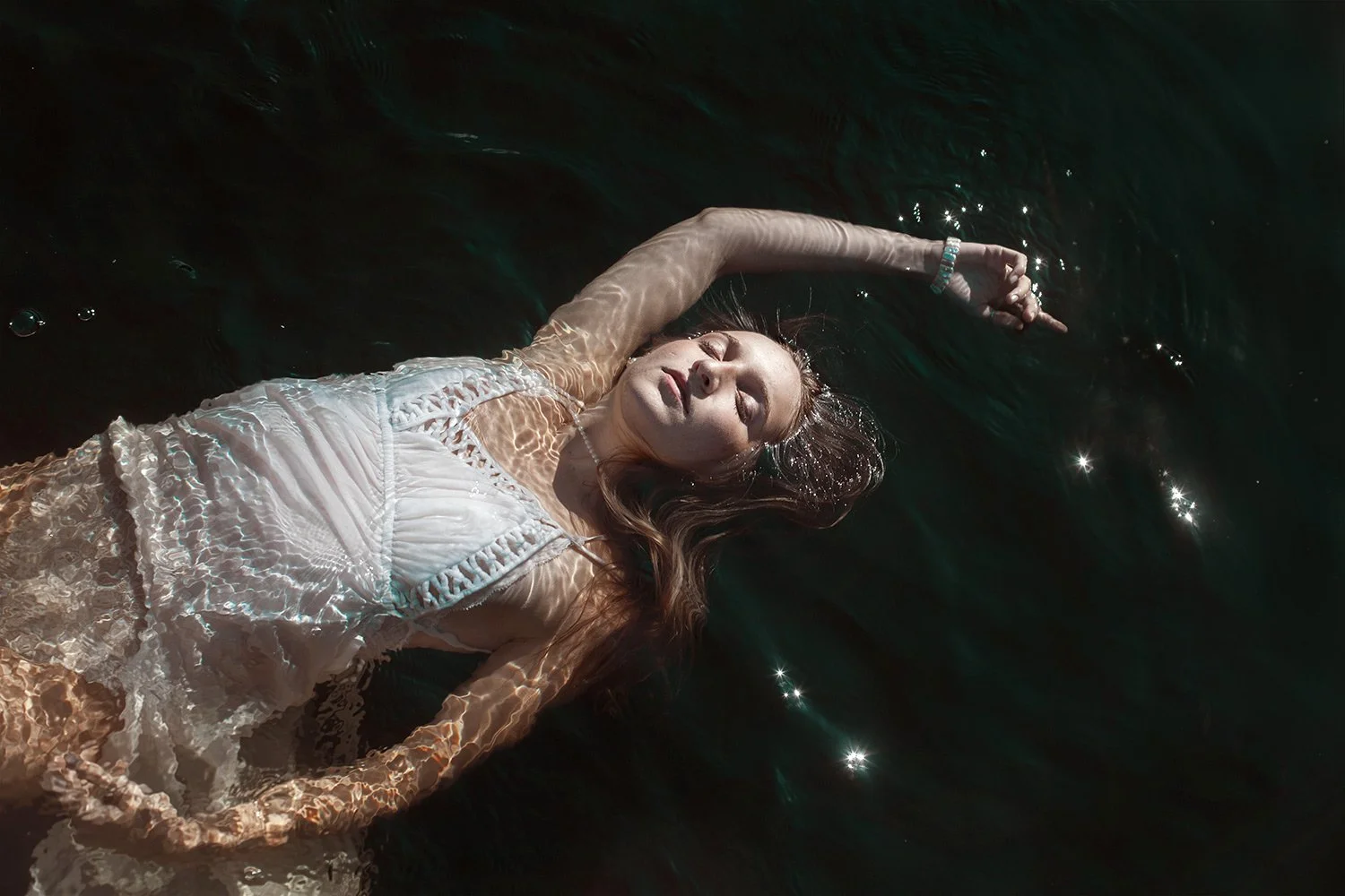 A woman in a white dress floating on dark water with sunlight creating reflections on her skin and water surface.
