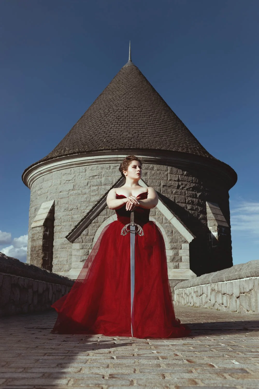 A woman in a red dress holding a sword stands in front of a round stone building with a conical roof.