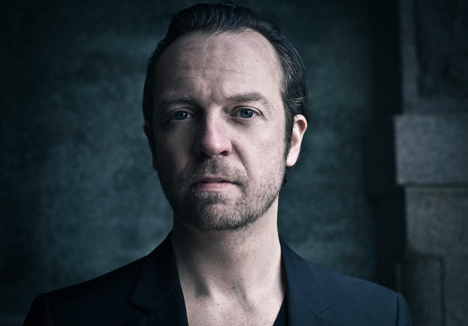 A portrait of a man with pale skin, blue eyes, and dark hair, wearing a black blazer, set against a dark background.
